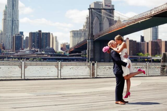 Paar küsst sich. Der Bräutigam hat die Braut hochgehoben in seinen Armen. Im Hintergrund die Brooklyn Bridge und die Skyline von New York.