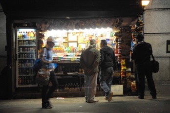 Blick auf Kiosk in Manhattan New York - Nächtliche Straßensezene - Drei Männer sind zum hellerleuchteten Kiosk gerichtet, einer geht vorbei. Mit Baseballmütze, Rucksack und Tasche, so als käme er von seiner Arbeit.