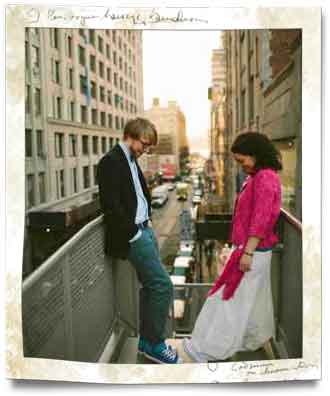 Hochzeitspaar in New York steht auf Brücke. Im Hintergrund Straßenschlucht, beide schauen auf ihre Schuhe und lächeln. Freuen sich über 
